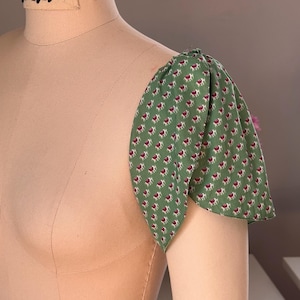 Puede incluir: Una tela verde con un patrón de elefantes pequeños blancos y rojos está fijada a un maniquí. La tela está fruncida en el hombro para crear una manga abullonada.