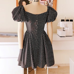 Puede incluir: Un vestido de lunares negros y blancos con mangas abullonadas y un corpi&ntilde;o ajustado. El vestido tiene un escote cuadrado y un cierre de botones en la parte delantera.