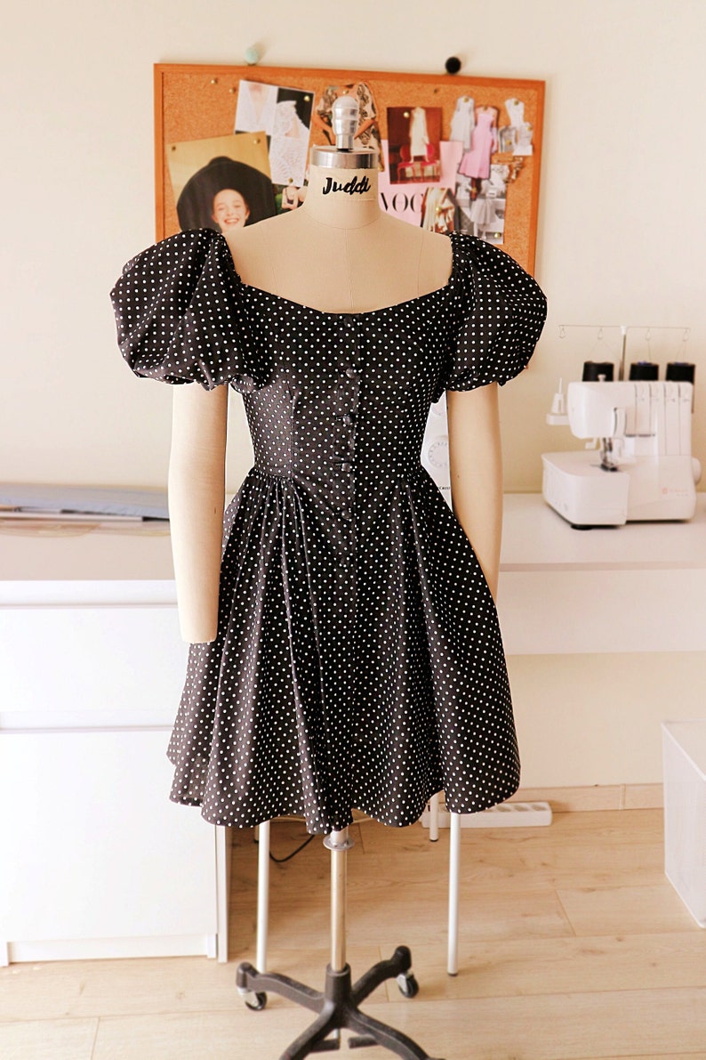 Puede incluir: Un vestido de lunares negros y blancos con mangas abullonadas y un corpi&ntilde;o ajustado. El vestido tiene un escote cuadrado y un cierre de botones en la parte delantera.