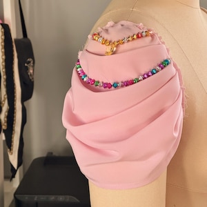 May include: A pink fabric sleeve with a gathered ruffle detail. The sleeve has two beaded necklaces on it, one gold and one multi-colored.