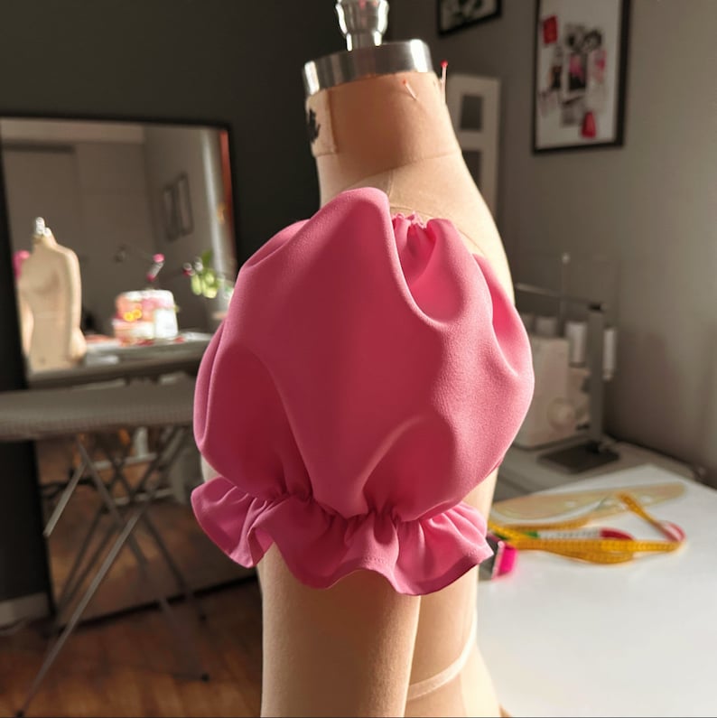 May include: A pink fabric puff sleeve with a ruffled edge. The sleeve is attached to a dress form.