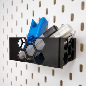 Op de afbeelding: Een zwarte zeshoekige wandbevestigde opbergbak met een blauw gereedschap en een witte kabel erin.