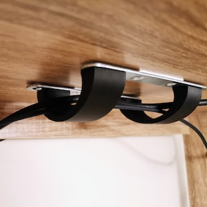 Könnte beinhalten: Nahaufnahme eines schwarzen Kabelmanagementsystems, das an der Unterseite eines Holzschreibtisches befestigt ist. Das System verfügt über zwei schwarze Clips, die die Kabel an Ort und Stelle halten.