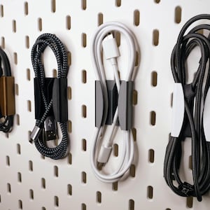 Op de afbeelding: Een verzameling oplaadkabels, elk vastgezet met een houder, wordt getoond tegen een witte pegboard. De kabels variëren in kleur, waaronder zwart, wit en een zwart-grijs gevlochten ontwerp. De houders zijn in tinten zwart, grijs en brons.