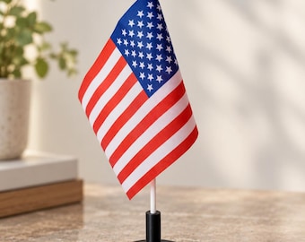 Puede incluir: Una pequeña bandera de escritorio de los Estados Unidos de América con las icónicas franjas rojas, blancas y azules y un cantón azul con estrellas blancas. La bandera de tela está unida a un poste blanco con un remate puntiagudo sobre una base negra.