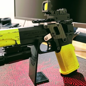 May include: A black and yellow pistol with a red dot sight and a torch attached. The pistol is on a black stand and has the text "OPS-M.R.P. CAL." on the side.