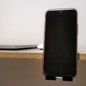 May include: A black smartphone with a clear case is mounted on a desk with a laptop and a white mouse in the background.