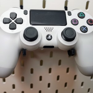 Puede incluir: Un mando de PlayStation 4 DualShock 4 blanco con botones y sticks analógicos negros. El mando está colgado en una pared de tablero perforado blanco.