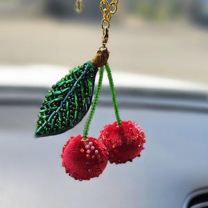 May include: A car charm featuring two red beaded cherries and a green beaded leaf, suspended from a gold-coloured chain. This decorative accessory adds a playful touch to a car's interior. The cherries are embellished with tiny red beads.
