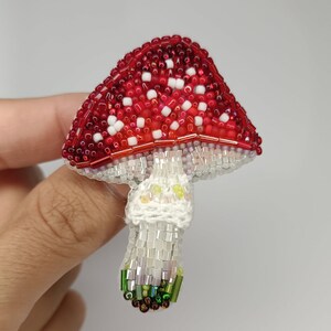 May include: A beaded mushroom brooch with a red and white cap, a white stem, and a green base.