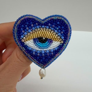 May include: A heart-shaped brooch crafted with blue, white, and gold beads, featuring a central eye design with a blue iris and black pupil. The perimeter is lined with metallic blue beads, and a single white pearl-like bead hangs from the bottom.