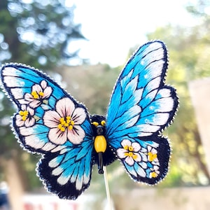 以下が含まれることがあります： 黄色いアクセントと白い花が付いた、青と白の刺繍の蝶のブローチ。蝶はピンに付いており、手で持たれています。