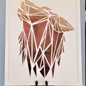 Puede incluir: Una pieza de arte geométrico de madera enmarcada que representa la cabeza de un lobo en un esquema de color marrón y blanco.