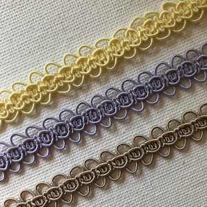 May include: Close-up of three rows of decorative trim in pastel colors. The top row is a pale yellow, the middle row is lavender, and the bottom row is a muted brown. Each row has a repeating looped design, perfect for sewing and craft projects.