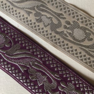 May include: Close-up of two decorative ribbons. One is a deep purple with silver floral and geometric patterns. The other is a light beige with similar silver designs. Both ribbons have a textured appearance and are suitable for crafts.