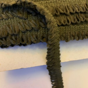 May include: A close-up of dark green fabric trim with a ruffled edge. The trim is folded in half and the ruffled edge is visible on both sides.