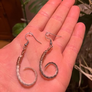 May include: A pair of unique earrings featuring a curled, gray and brown fur design. The fur is wrapped with copper wire and attached to silver-colored ear hooks. The earrings are displayed against a blurred background.