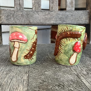 Op de afbeelding: Twee cilindrische keramische mokken met een lichtgroene glazuurafwerking. De mokken zijn voorzien van verhoogde, handgeschilderde ontwerpen van roodkapjespaddenstoelen met witte stippen en bruine, gesegmenteerde duizendpoten.