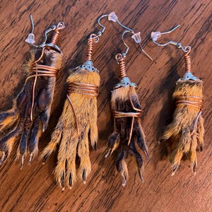May include: Four unique earrings featuring small animal paws. The paws are wrapped with copper wire and attached to silver-colored ear hooks. The fur on the paws is a mix of brown, tan, and yellow.