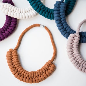 May include: Five different colored rope necklaces. The necklaces are made of thick, braided rope and are all different lengths. The colors are purple, white, teal, blue, and pink.