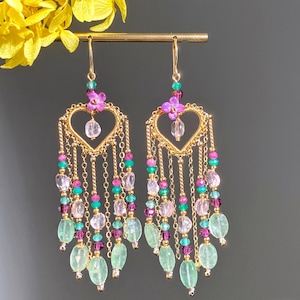 Peut inclure: Boucles d'oreilles pendantes dorées avec des cadres en forme de cœur et des chaînes en cascade. Les boucles d'oreilles sont ornées de perles roses, vertes et transparentes, et d'un petit détail floral. Elles sont suspendues à un crochet doré.