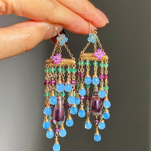 May include: Gold-toned chandelier earrings with cascading blue, green, and purple beads. The earrings feature a floral design with light blue and pink flowers. The earrings are held up by a hand.