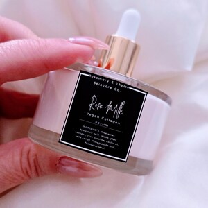 May include: A clear glass bottle of Rose Milk Vegan Collagen Serum with a white dropper and gold-colored top. The bottle has a pink label with black text that lists the ingredients. The product is from Rosemary & Thyme Skincare Co.