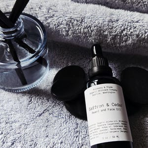May include: A black glass bottle of Saffron & Cedar Beard and Face Oil with a dropper, next to black stones and a glass diffuser with black reeds. The bottle is 2 oz / 50 mL.
