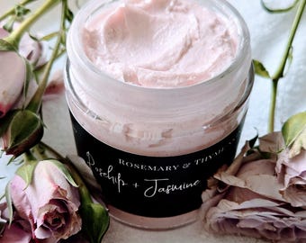 Puede incluir: Un frasco transparente lleno de crema rosa claro, con una etiqueta negra con texto blanco que dice Rosemary & Thyme y Rosehip + Jasmine. El frasco está rodeado por varias rosas de color violeta claro con tallos y hojas verdes.
