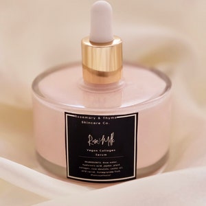 May include: A clear glass bottle filled with a pink serum, featuring a gold-colored collar and a white dropper. The bottle has a black label with white text that reads "Rose Milk Vegan Collagen Serum" and "Rosemary & Thyme Skincare Co."