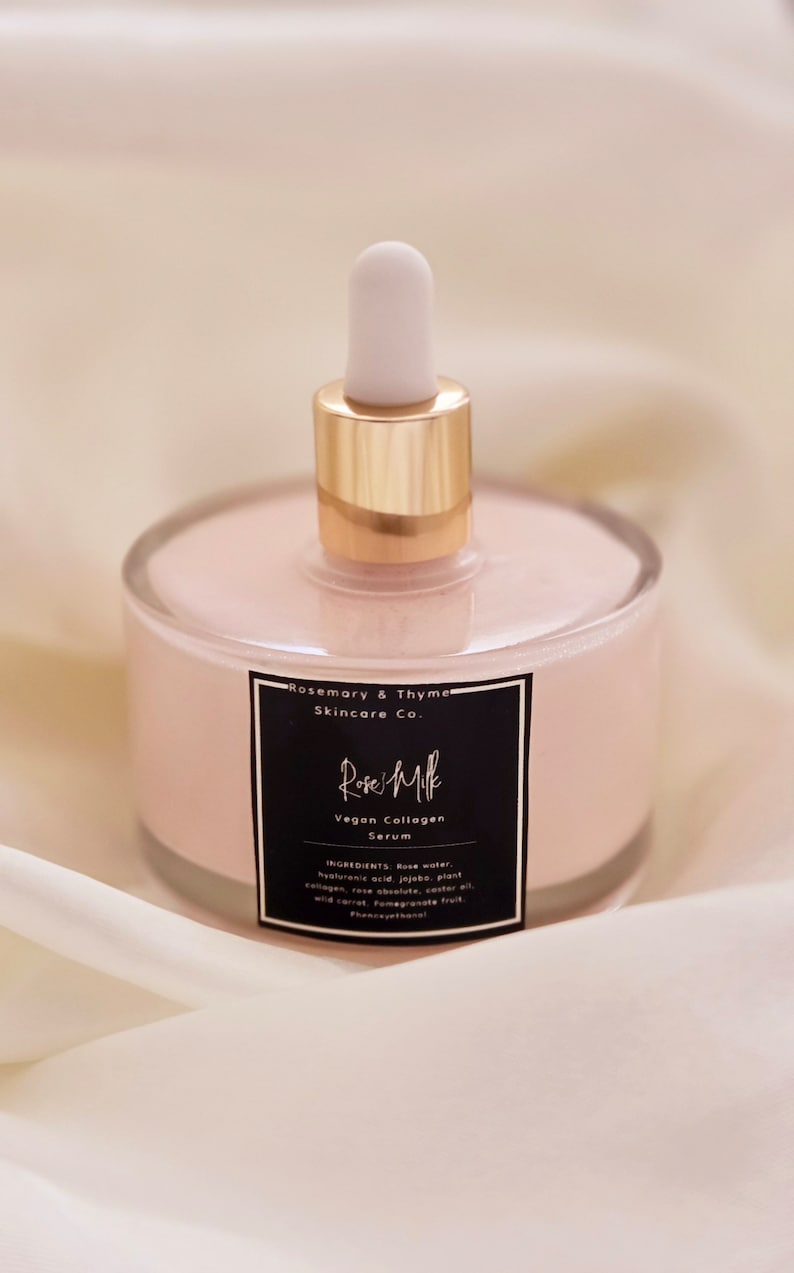 May include: A clear glass bottle filled with a pink serum, featuring a gold-colored collar and a white dropper. The bottle has a black label with white text that reads "Rose Milk Vegan Collagen Serum" and "Rosemary & Thyme Skincare Co."