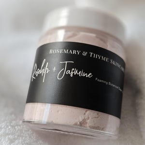 以下が含まれることがあります： 白い蓋が付いた、淡いピンク色の泡立つボタニカルシュガースクラブが入った透明なプラスチックジャー。黒いラベルには、白い文字で「Rosemary & Thyme Skincare」、「Rosehip + Jasmine」、「Foaming Botanical Sugar Scrub」と記載されています。