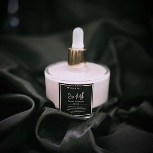 May include: A bottle of Rose Milk Vegan Collagen Serum with a white and gold dropper. The serum is in a clear glass bottle with a white base and a black label. The label includes the text "Rose Milk" and "Vegan Collagen Serum". The bottle sits on a black satin fabric.