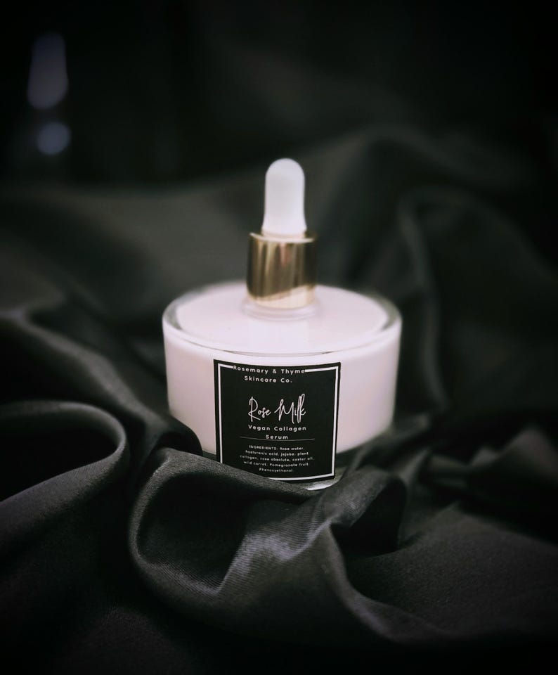 May include: A bottle of Rose Milk Vegan Collagen Serum with a white and gold dropper. The serum is in a clear glass bottle with a white base and a black label. The label includes the text "Rose Milk" and "Vegan Collagen Serum". The bottle sits on a black satin fabric.