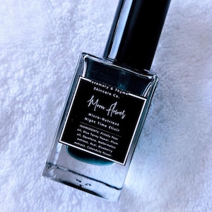 May include: A clear glass bottle of "Moon Flower" night time elixir, with a black cap and label. The label includes the text "Rosemary & Thyme Skincare Co." and ingredient information. The bottle is on a white, textured surface.