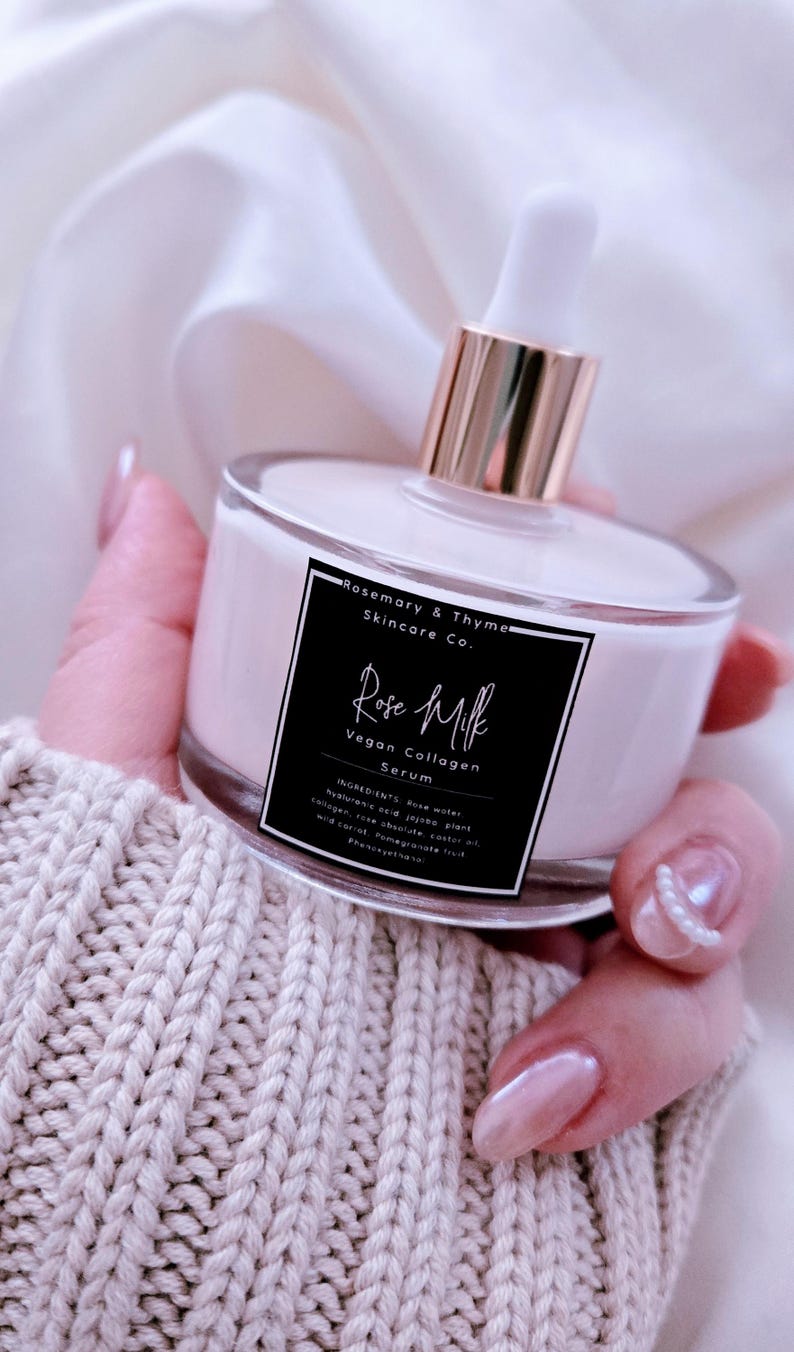 May include: A clear glass bottle of Rose Milk Vegan Collagen Serum by Rosemary & Thyme Skincare Co. The bottle has a white liquid inside and a gold-colored dropper. The label is black with white text. The bottle is held against a white background.