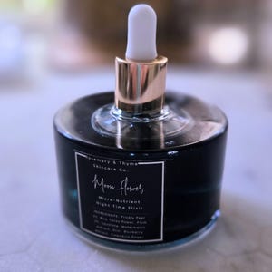 May include: A round, dark blue glass bottle of Rosemary & Thyme Skincare Co. Moon Flower Micro-Nutrient Night Time Elixir. The bottle features a gold-toned collar and a white rubber dropper top, with a black label listing ingredients in white text.