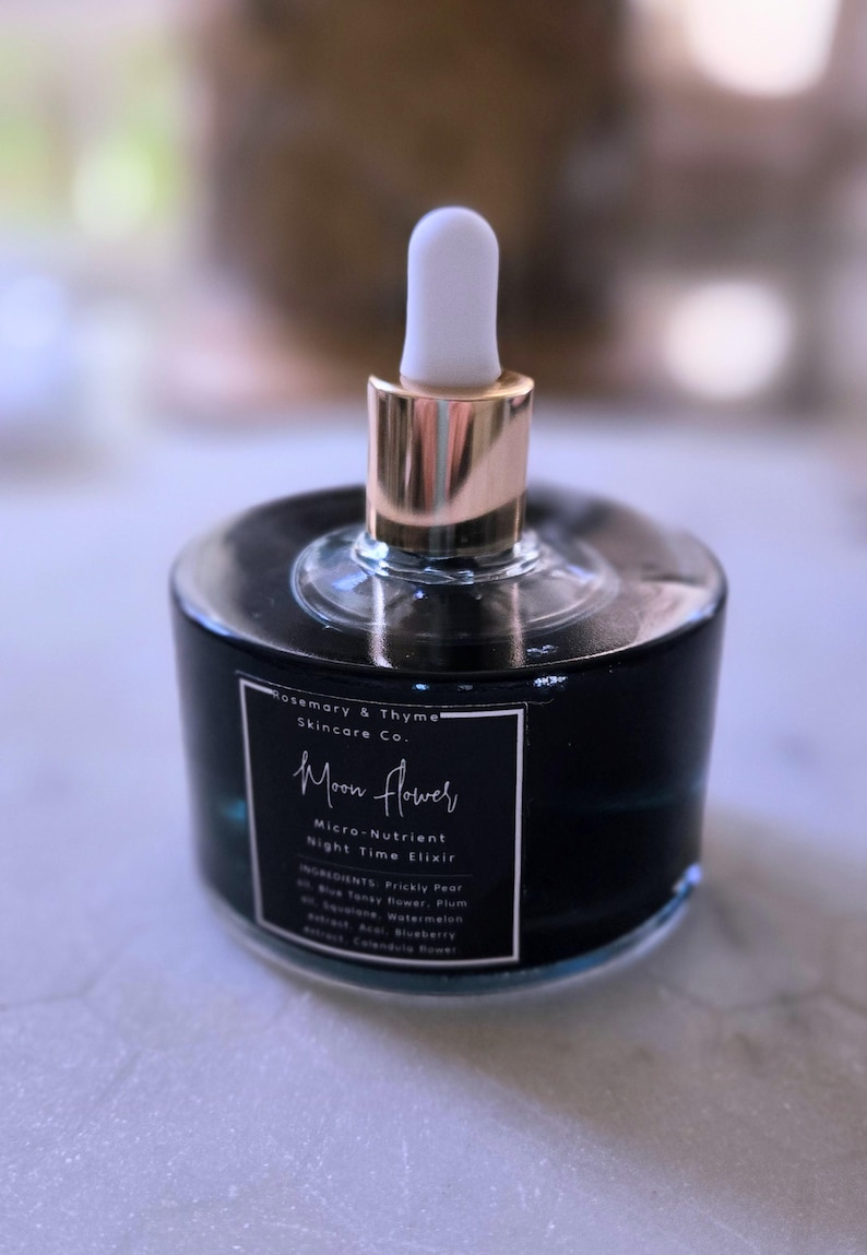 May include: A round, dark blue glass bottle of Rosemary & Thyme Skincare Co. Moon Flower Micro-Nutrient Night Time Elixir. The bottle features a gold-toned collar and a white rubber dropper top, with a black label listing ingredients in white text.