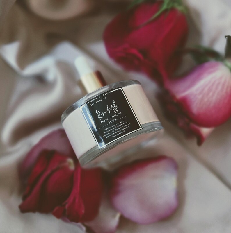 May include: A clear glass bottle of Rose Milk Vegan Collagen Serum with a white and black label, surrounded by red rose petals. The bottle has a gold-colored dropper and a light pink band around the middle.