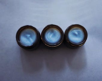 Puede incluir: Tres frascos pequeños y redondos de vidrio marrón oscuro dispuestos en una fila horizontal. Cada frasco está abierto y lleno de una crema o bálsamo de color azul claro con una superficie ligeramente texturizada y ondulada.