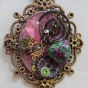 May include: Ornate pendant with a steampunk aesthetic. The pendant features a pink resin center with gears, chains, and a green and purple stone. The outer frame is a detailed bronze filigree.