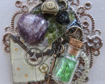Può includere: Un assemblaggio di tecniche miste con un telefono vintage, una pietra a forma di cuore viola e una piccola bottiglia di vetro con un tappo. Include anche parti di orologio, una chiave e una cornice decorativa. La tavolozza dei colori comprende bronzo, verde e viola.