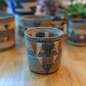 May include: A woven basket planter featuring a geometric pattern in shades of brown, beige, and grey. The planter is crafted from natural materials and has a distinctive, handcrafted look.
