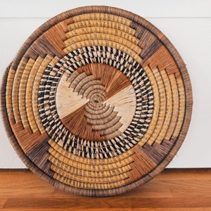 May include: A round woven wall hanging with a circular pattern of brown, black, and beige straw. The woven wall hanging is a decorative piece for a home.
