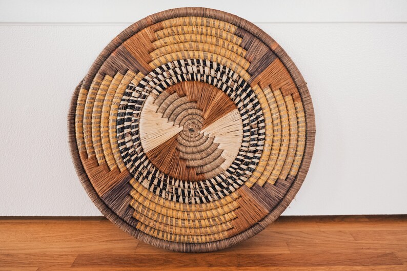 May include: A round woven wall hanging with a circular pattern of brown, black, and beige straw. The woven wall hanging is a decorative piece for a home.