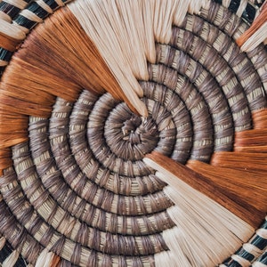 May include: Close-up of a woven basket with a circular pattern. The basket is made of natural materials in shades of brown, beige, and black.