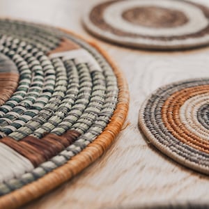 May include: Close-up of four woven round placemats in various shades of brown, beige, and green. The placemats are made of natural materials and have a textured surface.