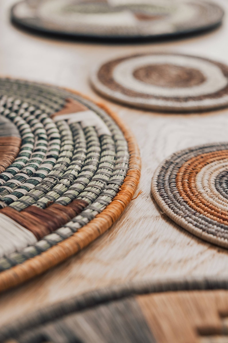 May include: Close-up of four woven round placemats in various shades of brown, beige, and green. The placemats are made of natural materials and have a textured surface.