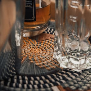 May include: A clear glass bottle with a brown liquid inside, sitting on a woven coaster with a black and white pattern. The bottle has a black label with white text that reads "at the that d, we artisan and um uses bose use with round strong in, vanilla leaves that you piced rum! le story at: distillery.com".