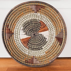 May include: A round woven wall hanging with a circular pattern in shades of brown, green, and white.